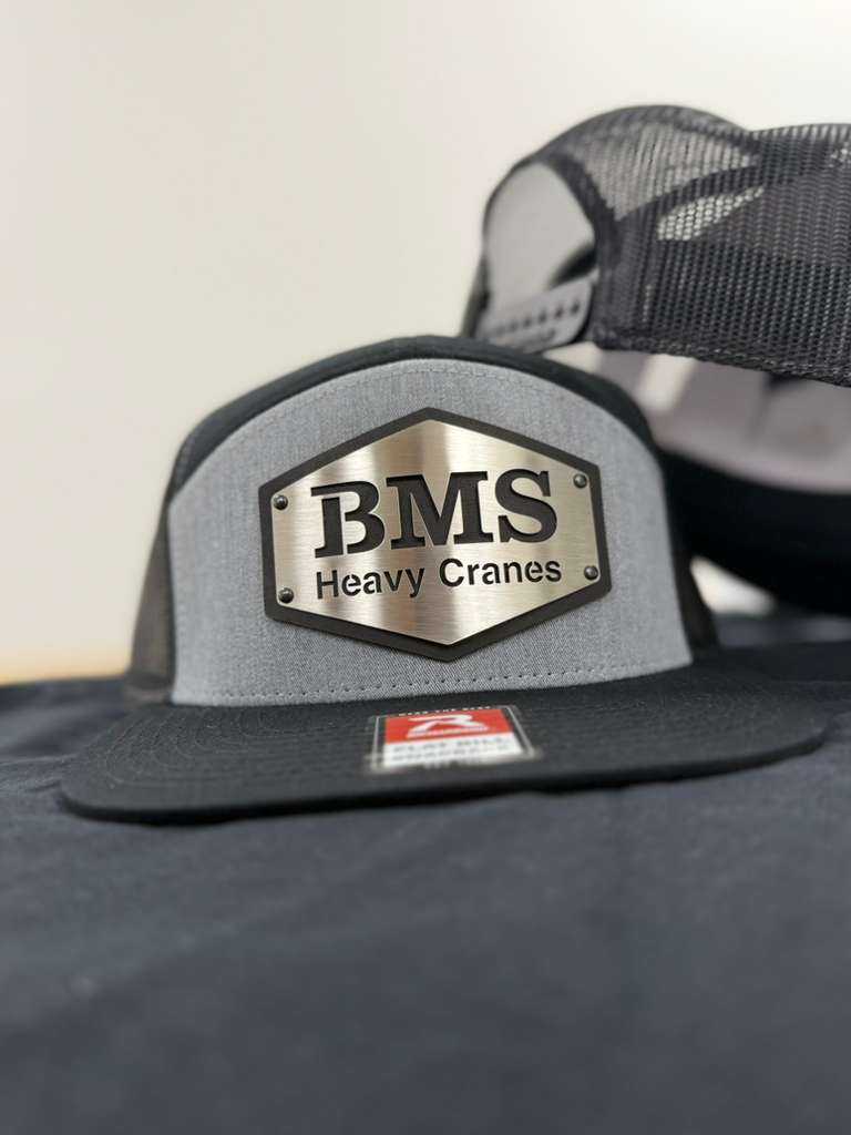 Close-up of BMS Black/Grey/Steel Trucker Hat featuring steel logo plate and mesh back, part of Job-Site Series workwear collection.