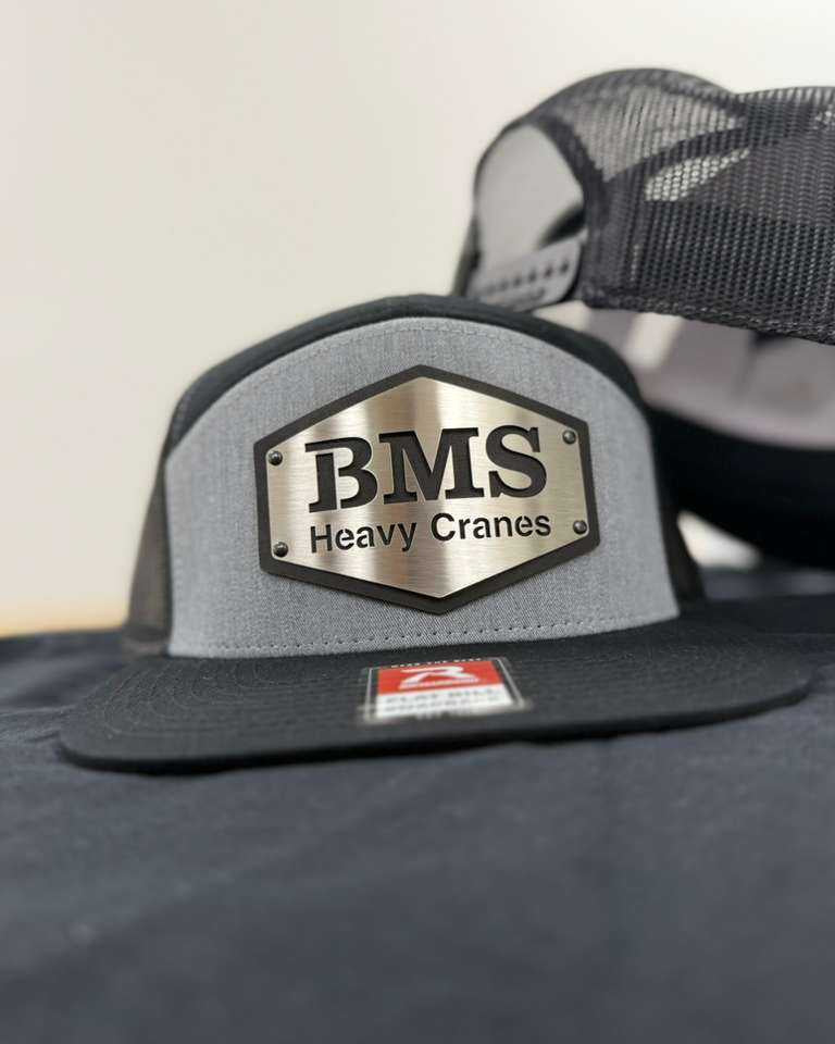 Close-up of BMS Black/Grey/Steel Trucker Hat featuring steel logo plate and mesh back, part of Job-Site Series workwear collection.