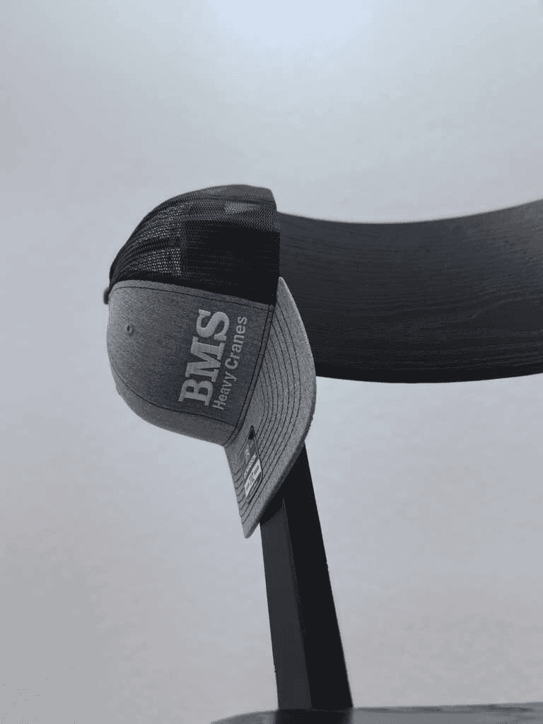 BMS Black/Grey Trucker Hat from Job-Site Series hanging on chair, showing mesh back and structured front panel with logo.