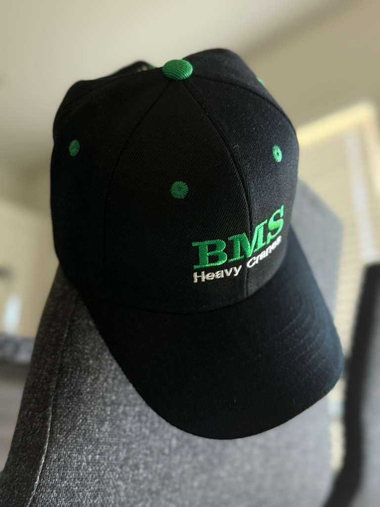 BMS Job-Site Series Black/Green Trucker Hat hanging on a chair, highlighting structured crown and green ventilation eyelets.