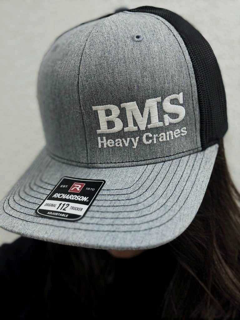 BMS Black/Grey Trucker Hat with mesh back and embroidered 'BMS Heavy Cranes' logo, worn on head, Richardson 112 frame visible.