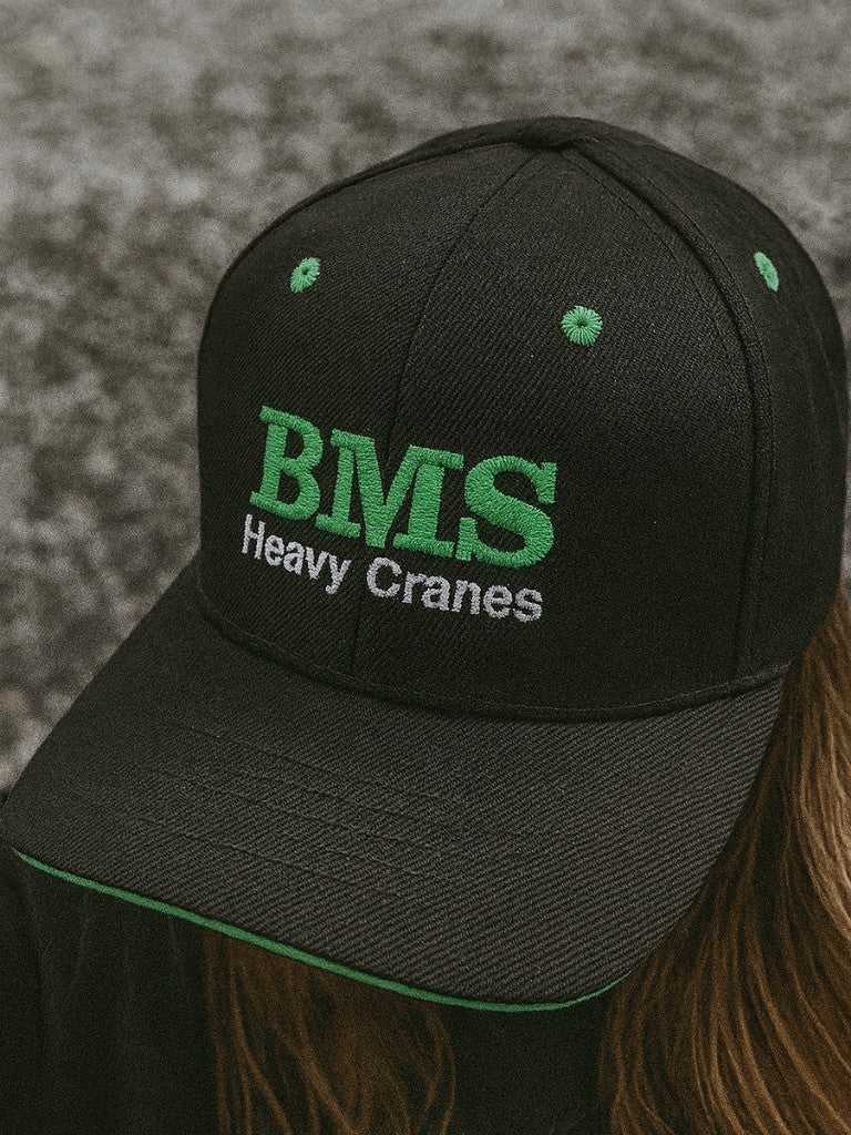 BMS Black/Green Trucker Hat with full back design, featuring green embroidered 'BMS Heavy Cranes' logo, ideal for job-site workwear.